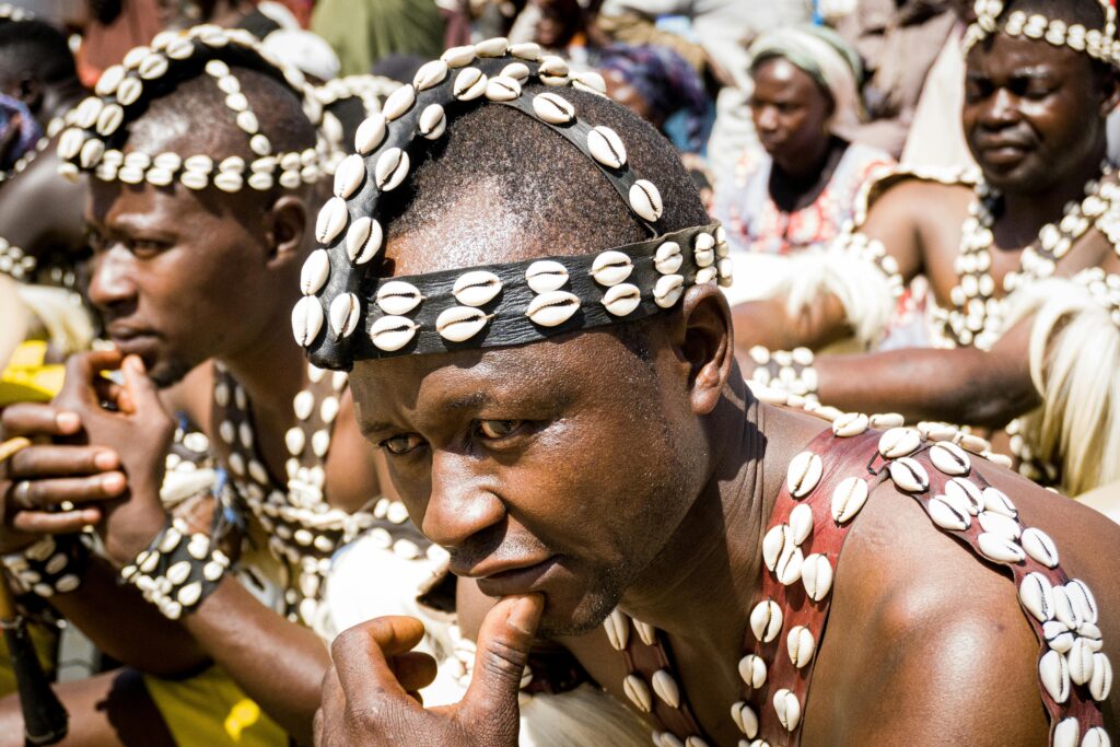 Men in traditional African attire wearing cowrie shell accessories, participating in a cultural dance.